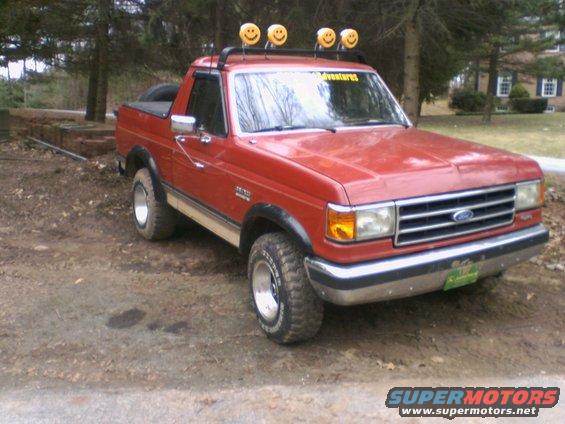 1989 Ford Bronco with smiley face lights on roof rack, parked on dirt