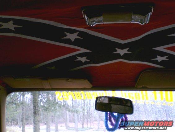 Inside a vehicle, looking up at a Confederate flag headliner. Sun visors are down. Rearview mirror hangs with blue and purple fuzzy things. Trees visible through the windshield