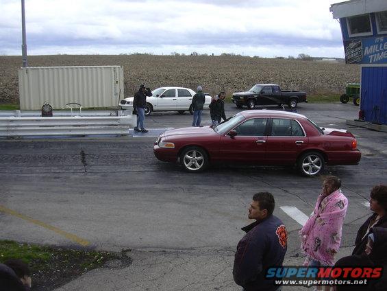 Maroon sedan and white sedan on a drag strip with people watching. Black pickup truck in background