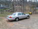 2004 Ford Crown Victoria - A silver 2004 Ford Crown Victoria is parked in a wooded area with houses in the background.
