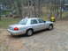 2004 Ford Crown Victoria - A silver sedan is parked outdoors on a gravel surface, with trees and houses in the background.