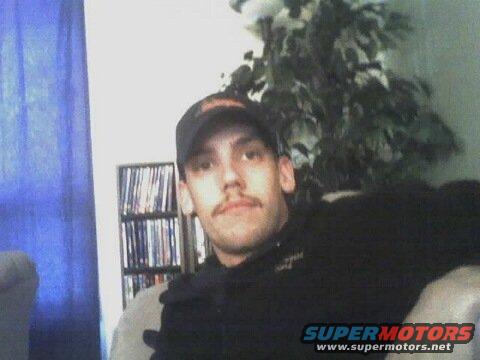 Man in black hoodie and baseball cap sits indoors, bookshelf behind him