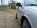 2004 Ford Crown Victoria - The side of a silver sedan is visible, parked on a gravel surface with trees and a body of water in the distance.