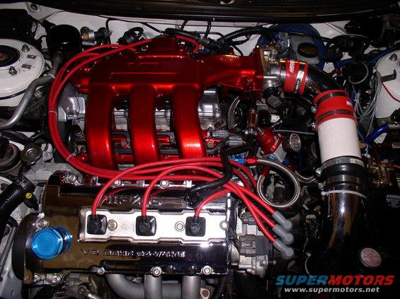 Engine bay with red intake manifold, chrome valve cover reading "V6 DOHC 24 VALVE", and red spark plug wires
