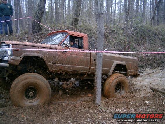 Mud-covered 1979 Jeep J-10 pickup truck with orange cab, driving through mud and trees