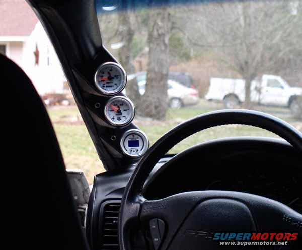 Interior view of a car's dashboard with three gauges mounted on the A-pillar and a steering wheel. Text 'SUPERMOTORS' visible on steering wheel