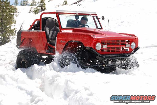 Red first-generation Ford Bronco with large tires driving through deep snow. Driver visible