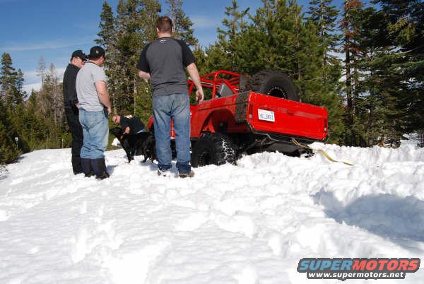 Red off-road vehicle with roll cage stuck in deep snow, people and a dog nearby
