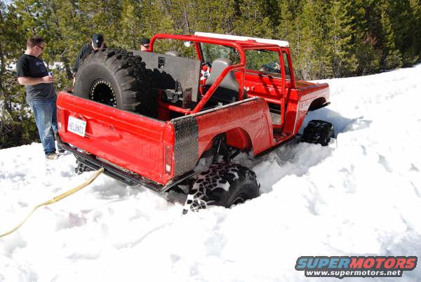 Red off-road vehicle stuck in snow, rear view, large spare tire in bed, tow strap attached