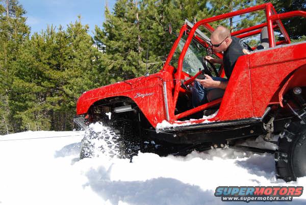 Red 1989 Ford Bronco with roll cage driving through snow, driver visible