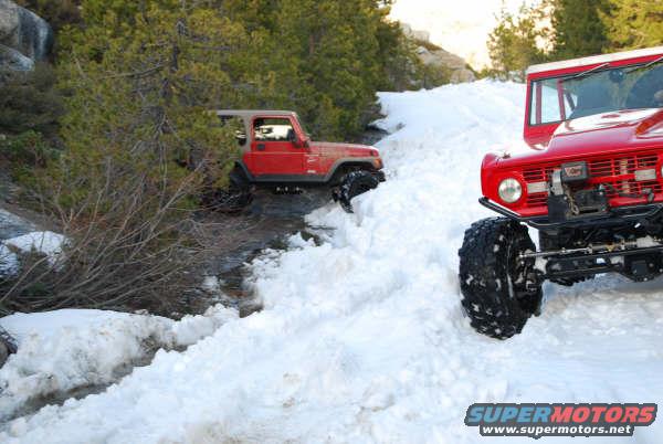 Red Jeep Wrangler and red Ford Bronco driving in snow, one on a snowy bank, the other in a snowy ditch