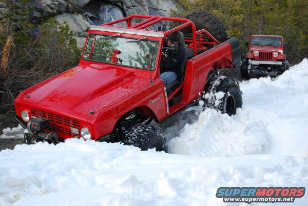 Red 1966 Ford Bronco off-roading in deep snow, with a Jeep Wrangler behind