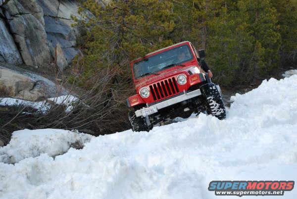 Red Jeep Wrangler climbing a snowy hill with trees and rocks in the background. License plate visible