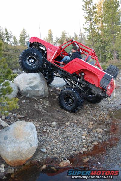 Red off-road SUV with roll cage climbing over rocks. Driver visible inside