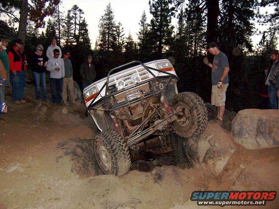 White SUV with roll cage going over rocks, people watching
