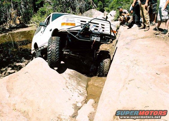 White off-road truck with custom bumper and winch going through a dirt ditch. People watch from the side