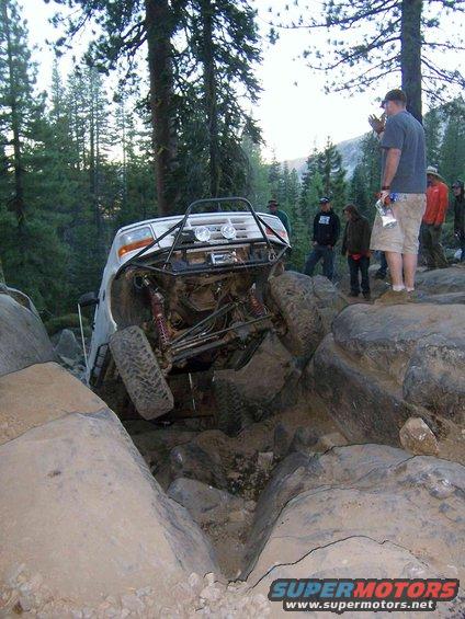 White off-road SUV with roll cage stuck between large rocks on a trail. People watch from the side
