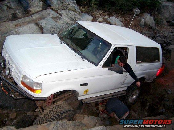 White 1995 Ford Bronco stuck on rocks while off-roading, with people trying to help