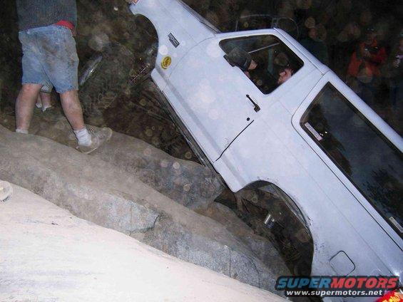 White Ford Bronco tilted on rocks, driver visible inside