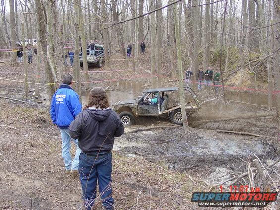 Off-road buggy driving through a muddy obstacle course in a wooded area. Spectators watch from the side