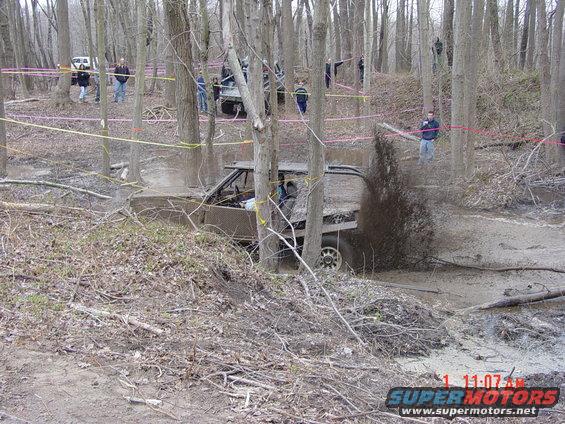 Mud-covered off-road truck with open doors driving through a muddy ditch, splashing water