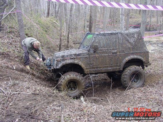 Mud-covered off-road vehicle stuck in mud with a person attempting to attach a winch line