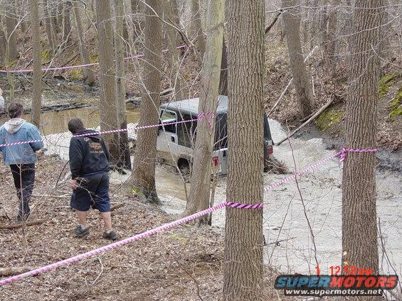 Silver Jeep Wrangler stuck in mud on a wooded trail, with two people observing