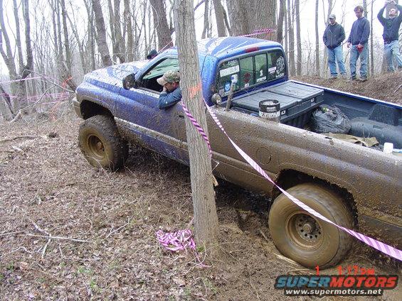 Blue pickup truck stuck in mud on a wooded trail, driver looking out window. Pink ribbon marks course