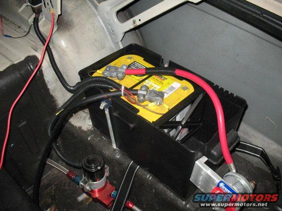 Car battery in a black box with red and black cables. Some fuel lines and a pump are visible below