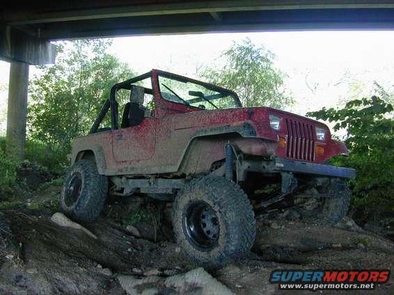 Red Jeep Wrangler on a muddy trail under a bridge