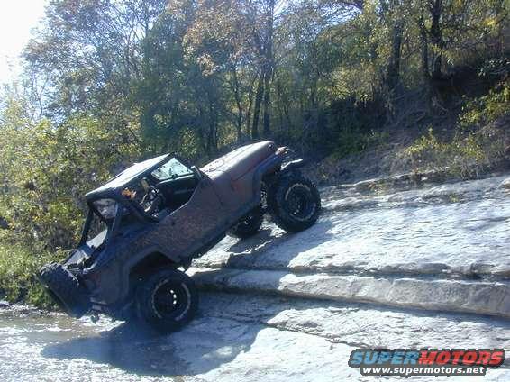 Brown off-road vehicle with large tires climbing over rocks near water