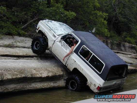 White off-road SUV with a black top, tilted on rocks in water, driver visible inside