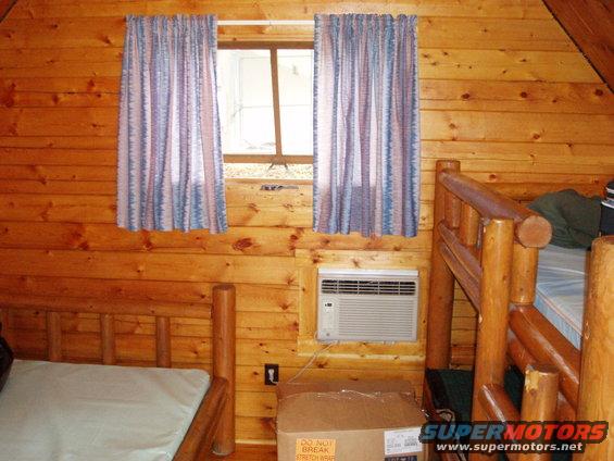 Interior of a log cabin room showing bunk beds, a window with curtains, and an air conditioning unit