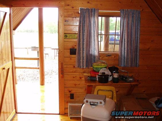 View from inside a wood-paneled cabin looking out an open door and through a window. A blue vehicle is visible outside