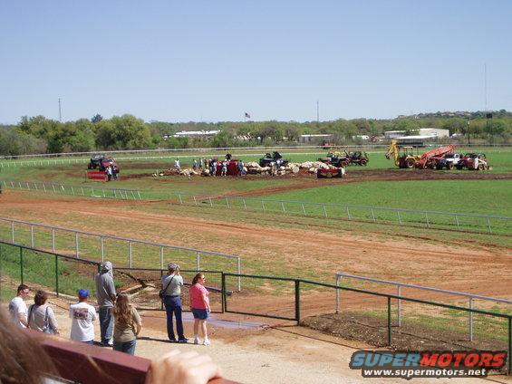 Several off-road vehicles and construction equipment are gathered on a dirt track with spectators watching