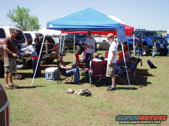 People hang out under tents at an outdoor event with several trucks visible in the background