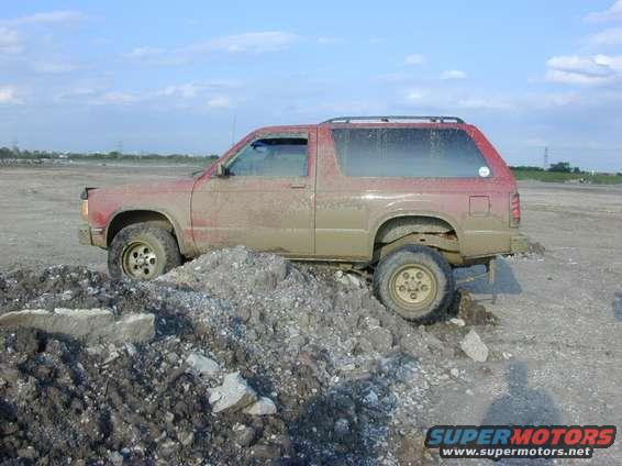 Mud-covered red SUV with a broken front fender, stuck on a pile of dirt