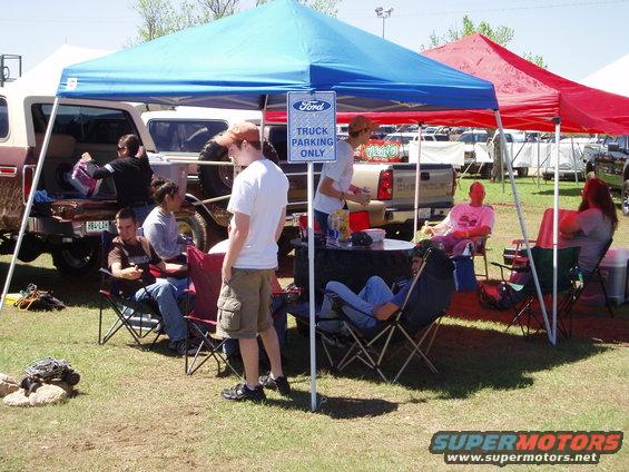 People sit in chairs under blue and red tents at an outdoor event; vehicles are parked in the background
