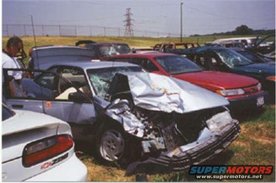 Wrecked silver sedan with crumpled front end, driver's door open, parked in a lot with other cars