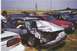 1996 Ford Bronco - A severely damaged silver sedan sits in a junkyard with its front end destroyed and driver's door ajar.