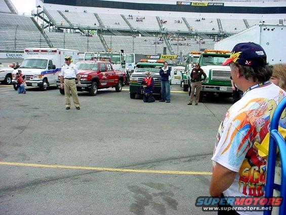 NASCAR pit area with tow trucks, an ambulance, and people standing around