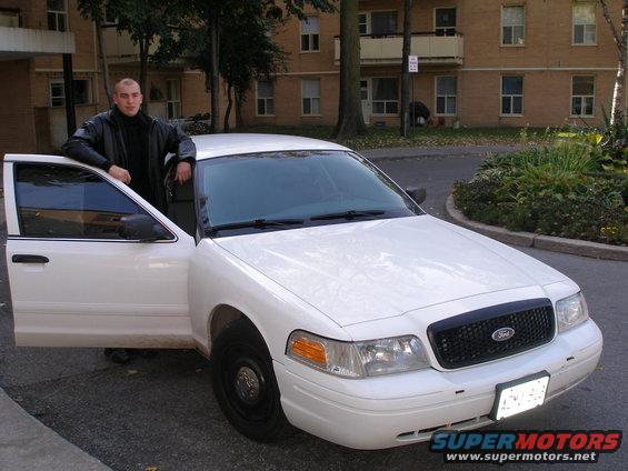 Man stands by a white Ford Crown Victoria with its driver's door open. The grille has a Ford logo
