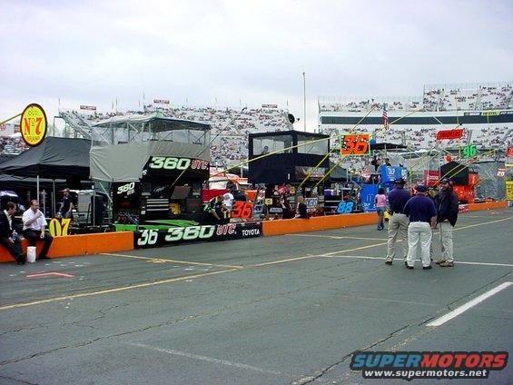NASCAR pit lane with race cars, crew members, and grandstands filled with spectators
