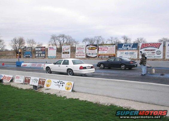 1998 Ford Crown Victoria and a dark sedan on a drag strip with advertising billboards in the background