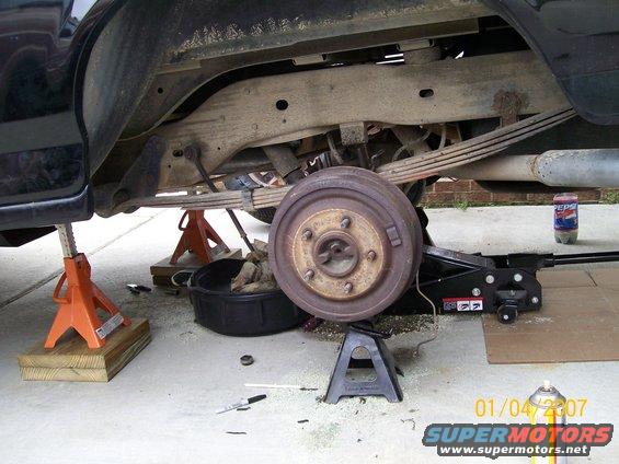 Vehicle rear axle assembly on jack stands, with brake drum visible. Oil drain pan and tools on the ground