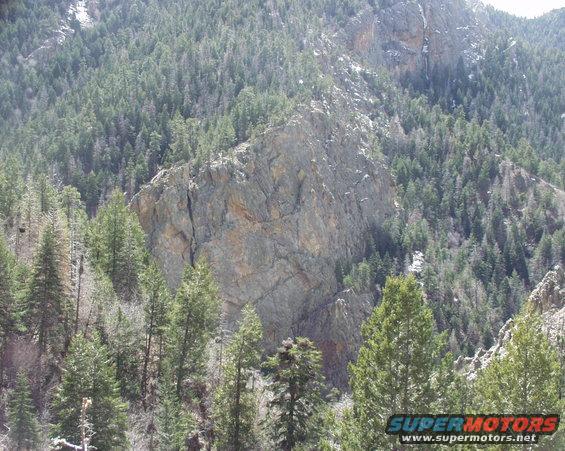 Mountain landscape with trees and a rocky cliff face. "SUPERMOTORS www.supermotors.net" text at bottom right