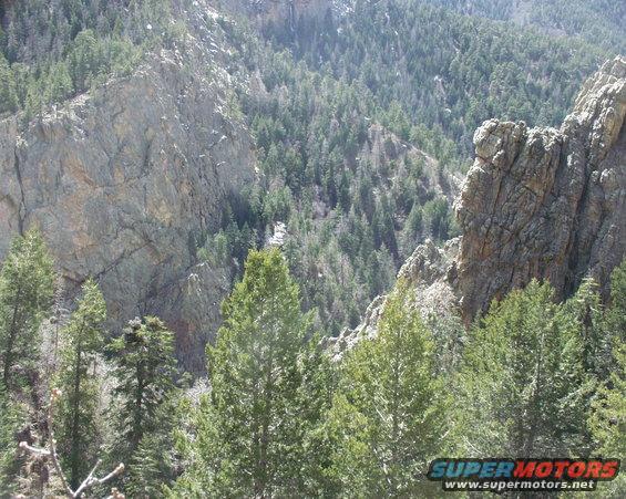 Mountain landscape with pine trees and rocky cliffs. SuperMotors.net logo in lower right