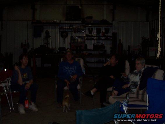 Four people sit in chairs in a dimly lit garage with a dog nearby