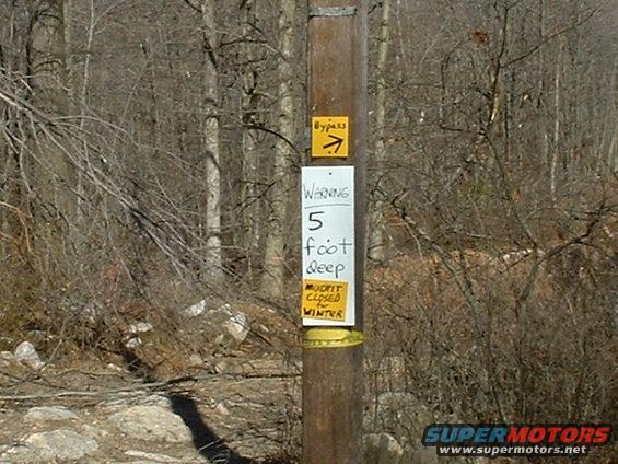 Wooden post with trail signs indicating a bypass and a warning about 5-foot deep mud, closed for winter
