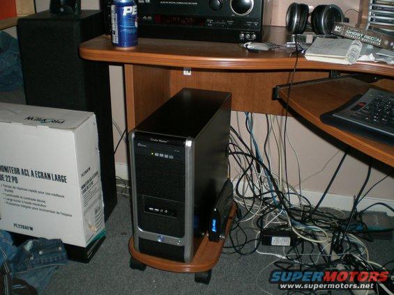 Computer tower on a wooden stand next to a large box, with a desk and tangled wires in the background